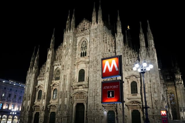 milan cathedral illuminated at night with a red metro sign in the foreground against the dark sky in a busy city square