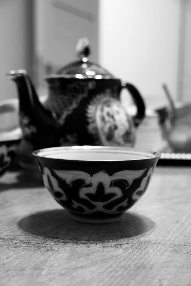 black and white close-up of a patterned ceramic teacup and teapot on a wooden table with soft indoor light and shallow focus