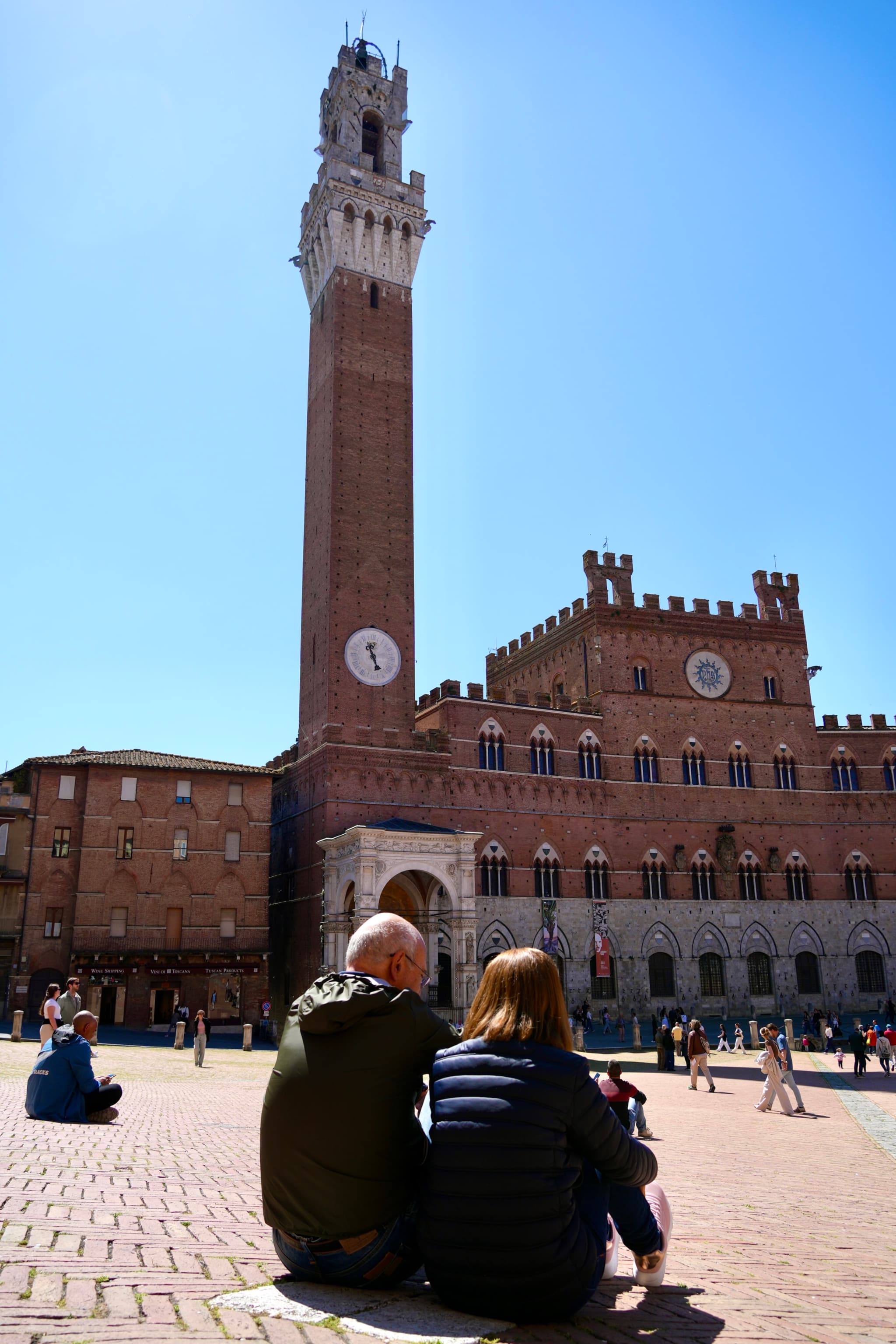 two people sit on the brick ground of piazza del campo in siena under bright sunlight facing the tall brick torre del mangia