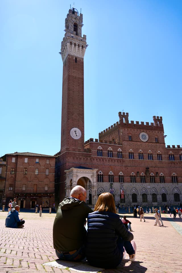 two people sit on the brick ground of piazza del campo in siena under bright sunlight facing the tall brick torre del mangia