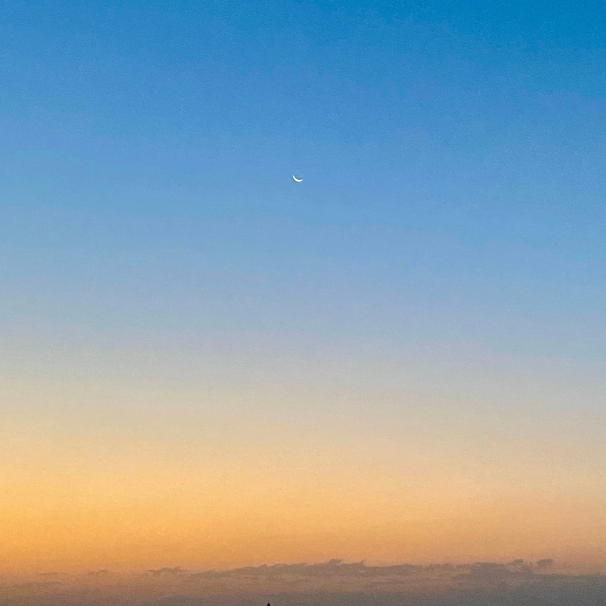 small crescent moon centered in a clear blue sky above a yellow horizon line during a quiet evening sunset in late summer