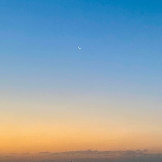small crescent moon centered in a clear blue sky above a yellow horizon line during a quiet evening sunset in late summer