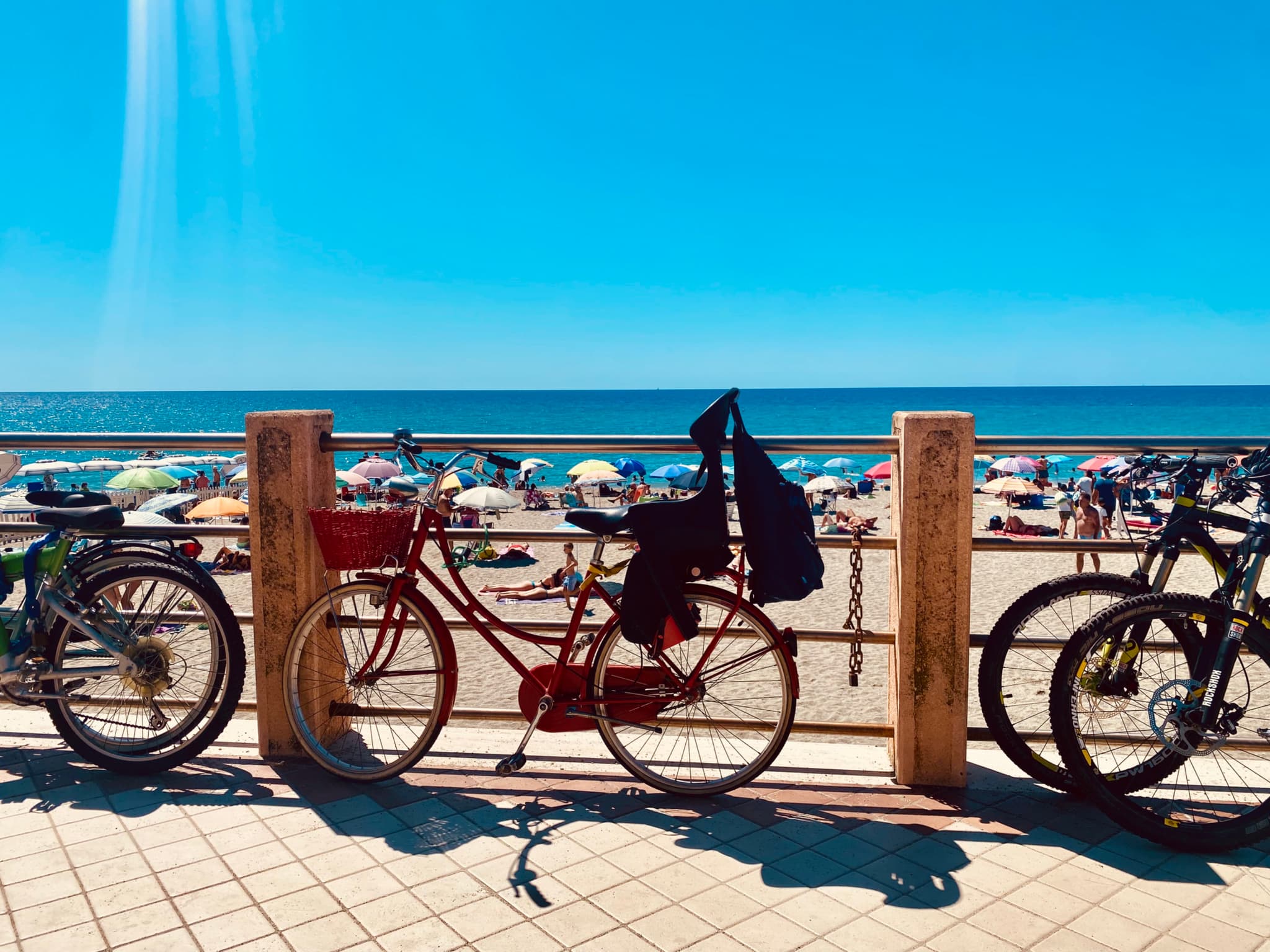 red bicycle parked on a sunny boardwalk overlooking a crowded beach with blue water under a clear sky during the day