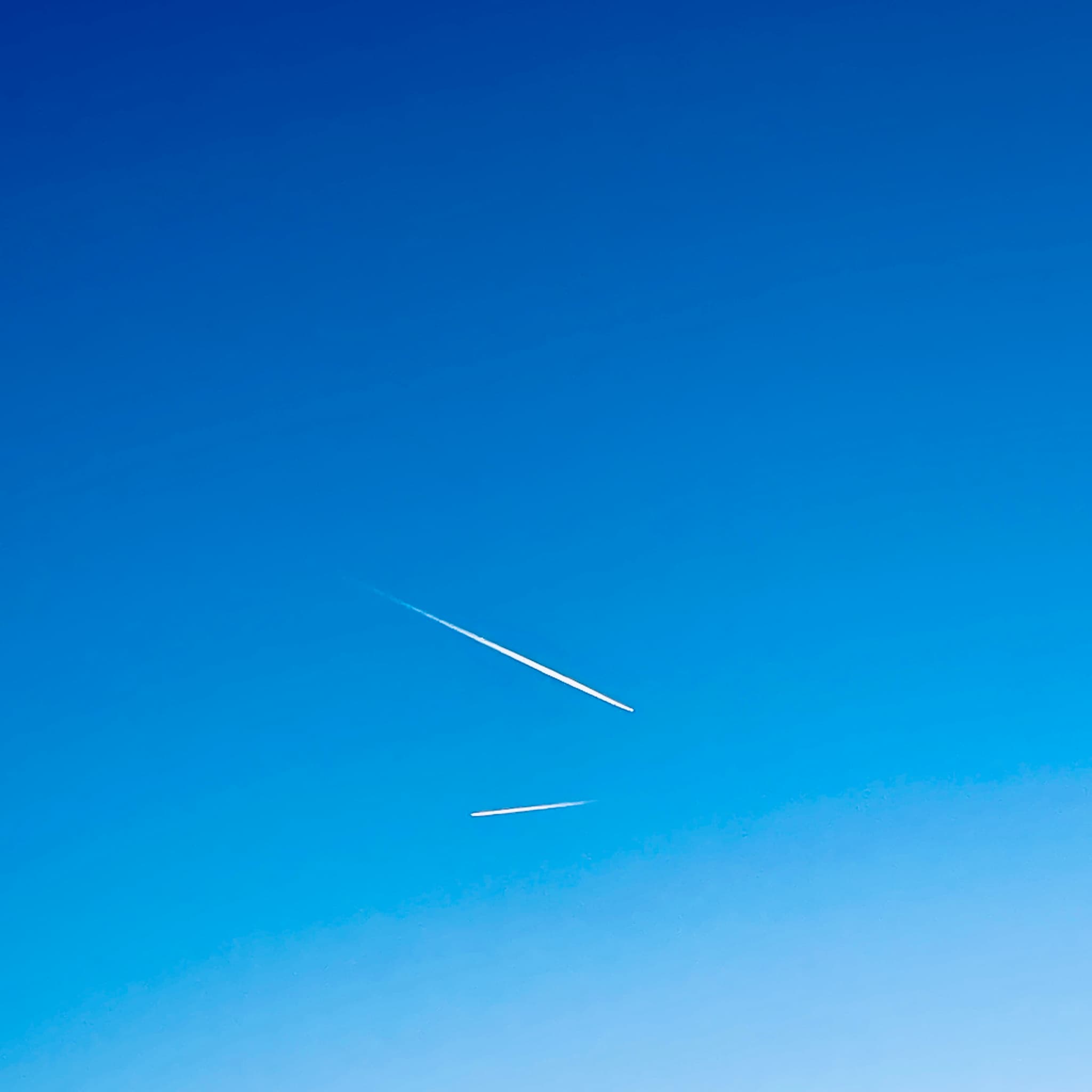 two white airplane contrails stretch across a clear blue sky during a bright day, creating thin lines against the open atmosphere
