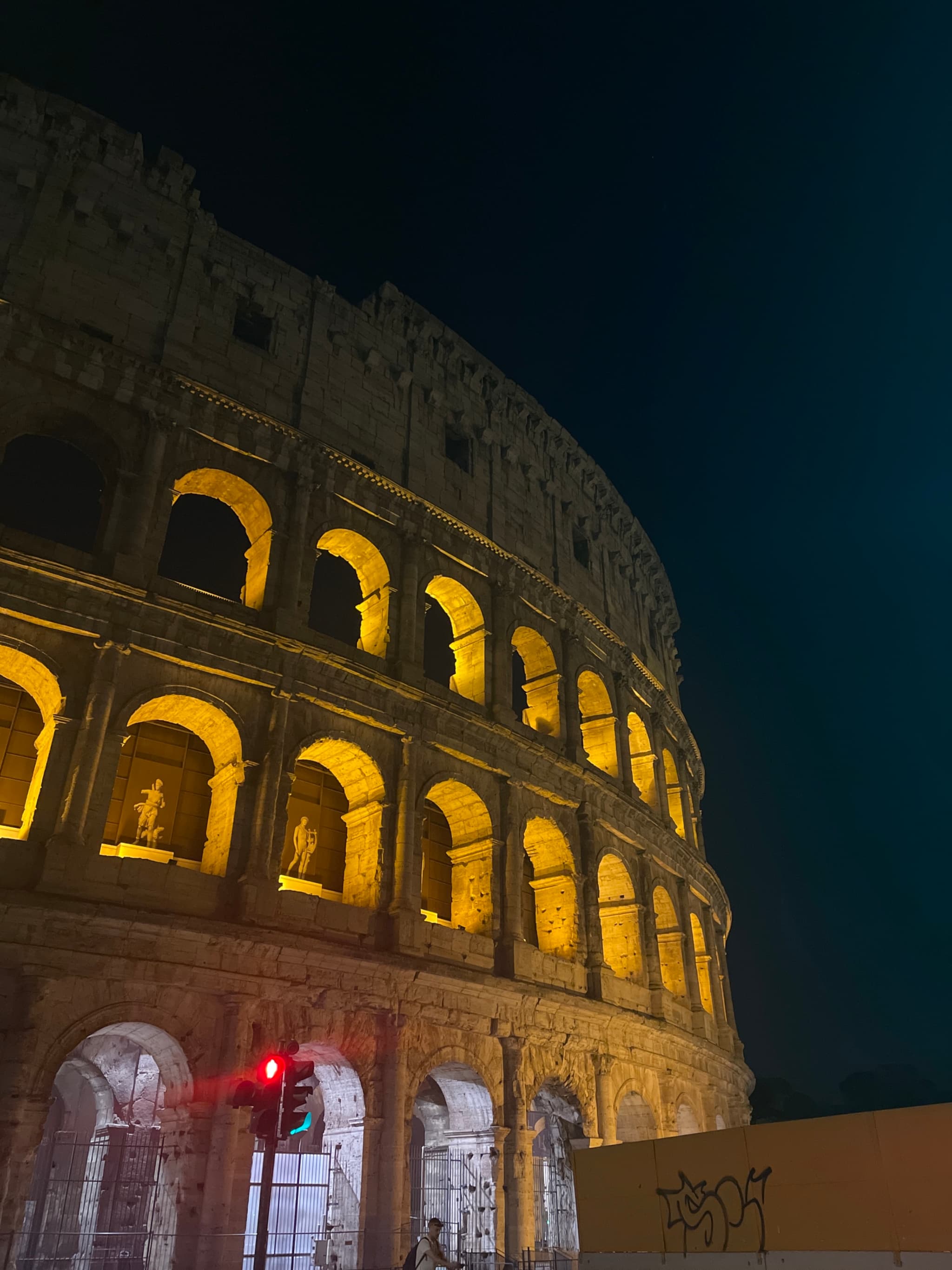 the colosseum in rome lit by yellow floodlights at night, showing stone arches against a dark sky with a traffic signal nearby