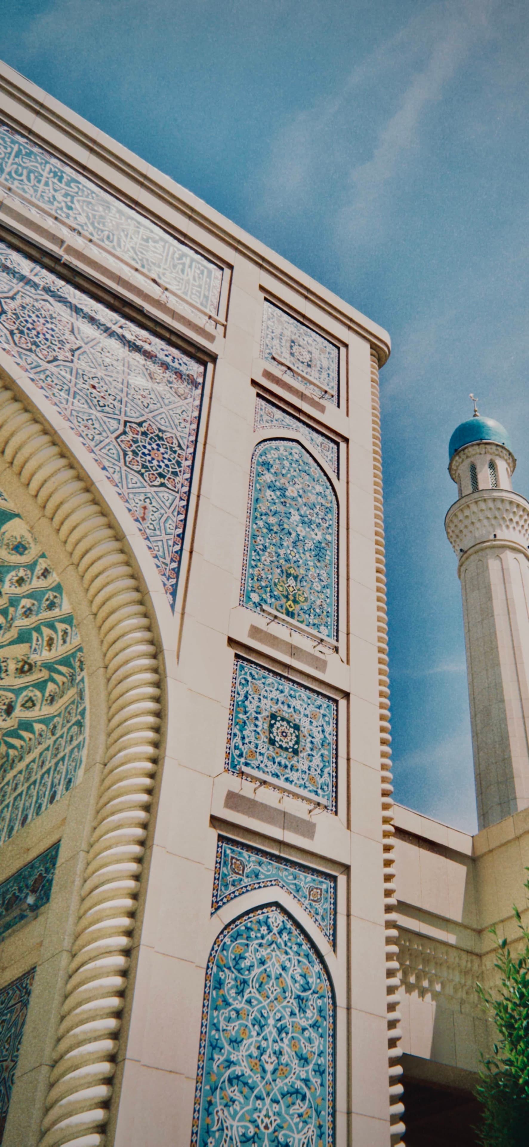 low angle view of a mosque archway with blue tile patterns and a tall minaret under a clear midday sky in daylight
