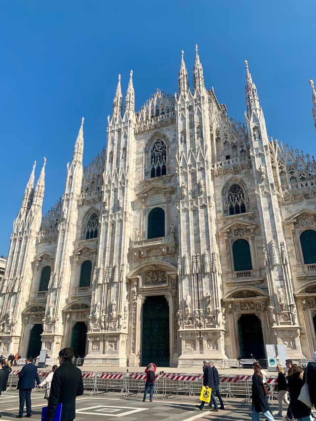white marble facade of milan cathedral under a clear blue sky with tourists walking across the paved square in bright daylight