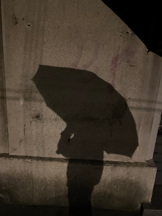 dark shadow of a person holding an umbrella cast onto a concrete wall at night under dim artificial street lighting