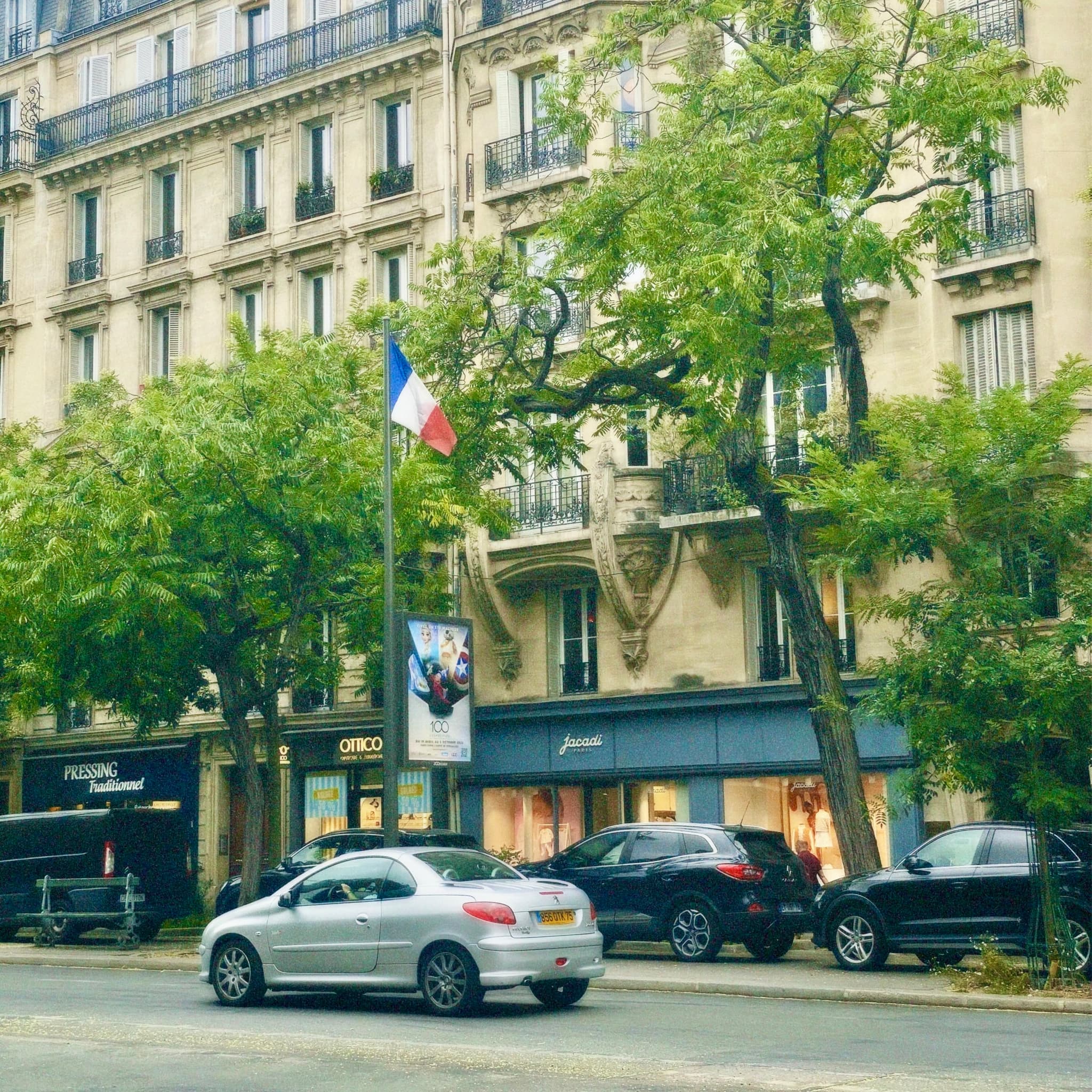 silver car parked on a parisian street in daylight with green trees, stone buildings, and a french flag under overcast sky