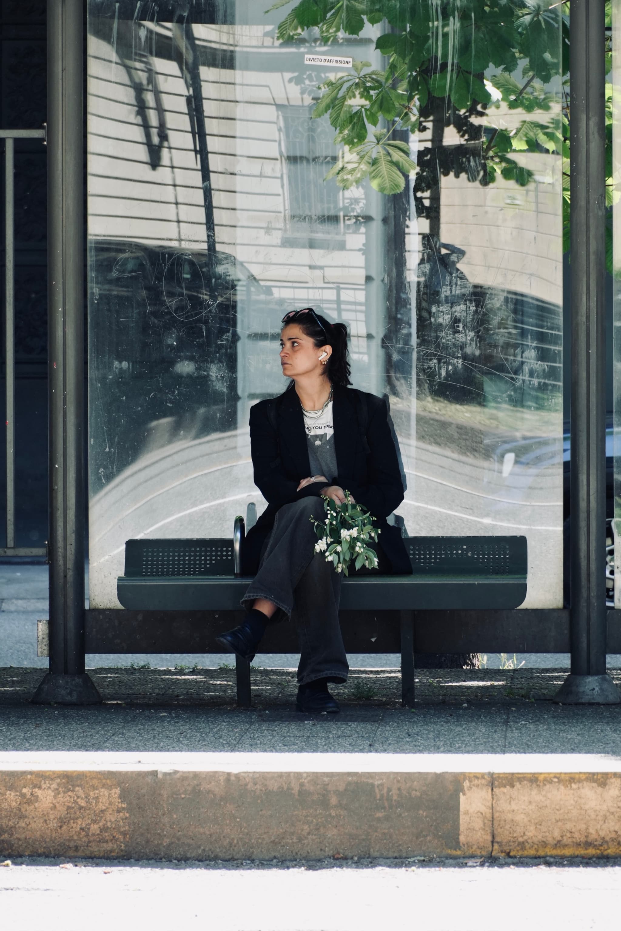 woman in a dark blazer sits on a glass bus stop bench holding flowers under bright daylight in an urban setting