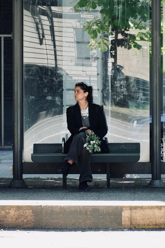 woman in a dark blazer sits on a glass bus stop bench holding flowers under bright daylight in an urban setting