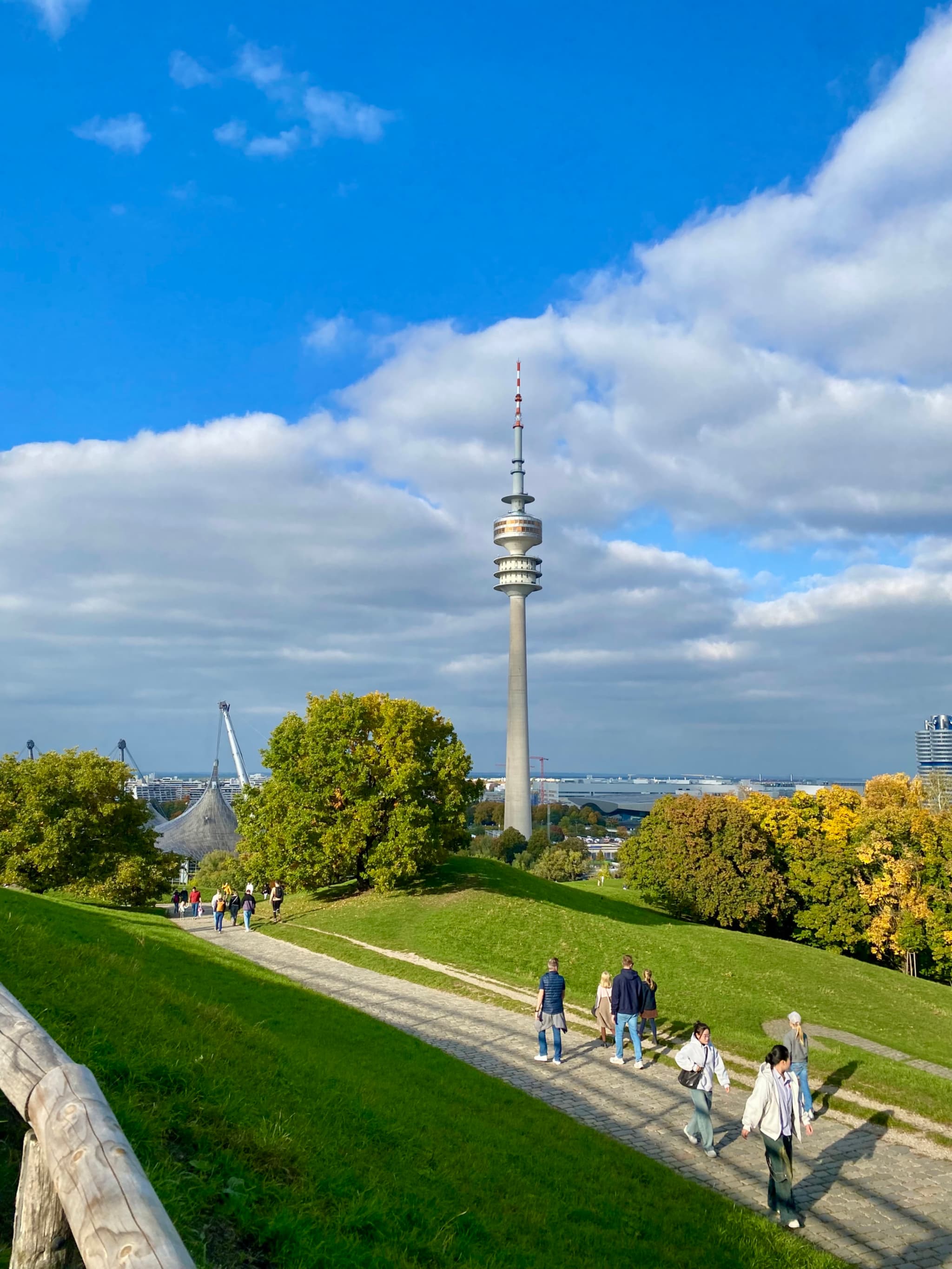 olympic tower stands above munich olympic park on a bright day with green hills, walking paths, and scattered white clouds overhead