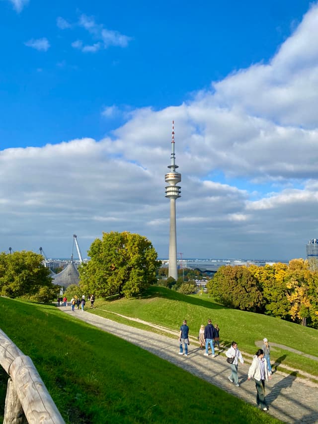 olympic tower stands above munich olympic park on a bright day with green hills, walking paths, and scattered white clouds overhead