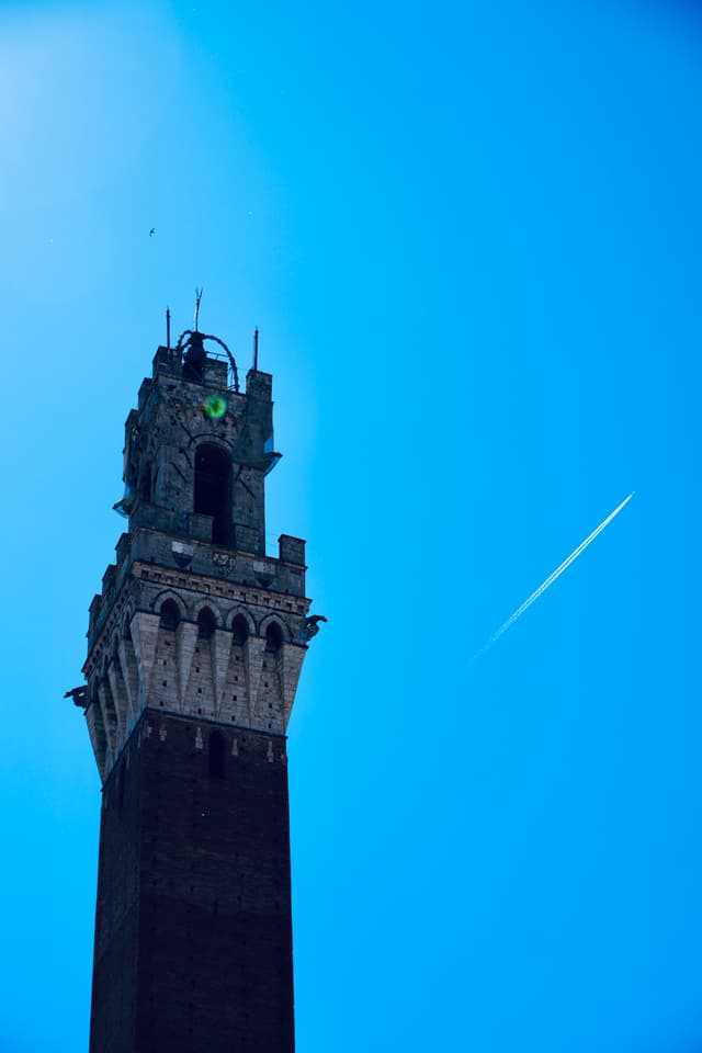 torre del mangia in siena against a clear blue sky with a white airplane contrail visible during the late afternoon light