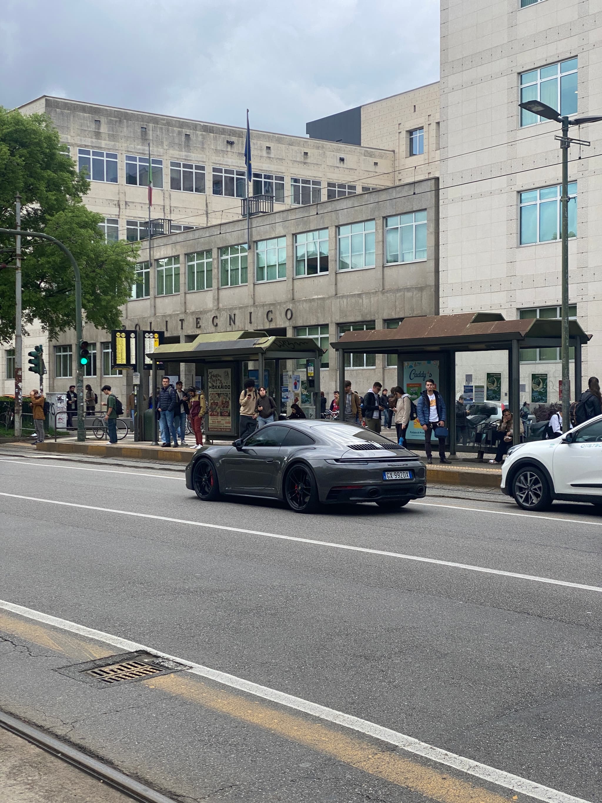 grey porsche driving past a crowded city bus stop on a cloudy afternoon with urban buildings and street traffic in the background
