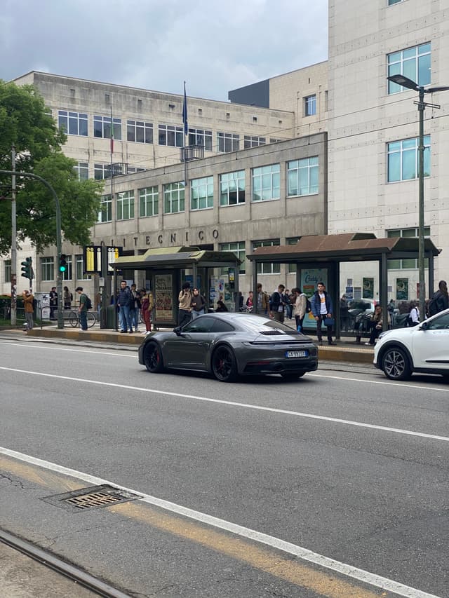 grey porsche driving past a crowded city bus stop on a cloudy afternoon with urban buildings and street traffic in the background