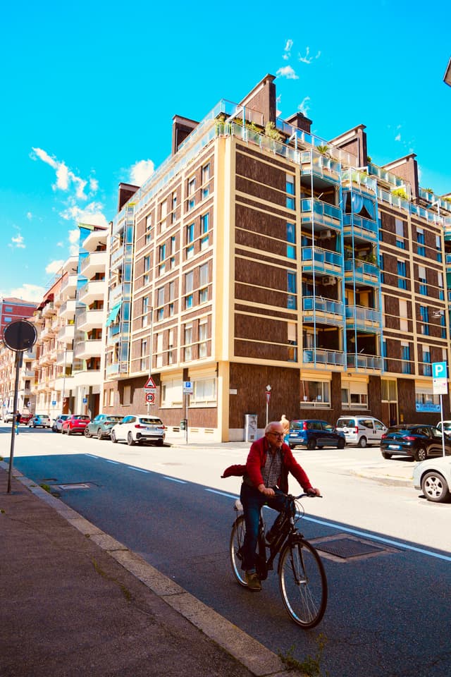 man in a red jacket rides a bicycle on a sunny city street past a large apartment building with glass balconies