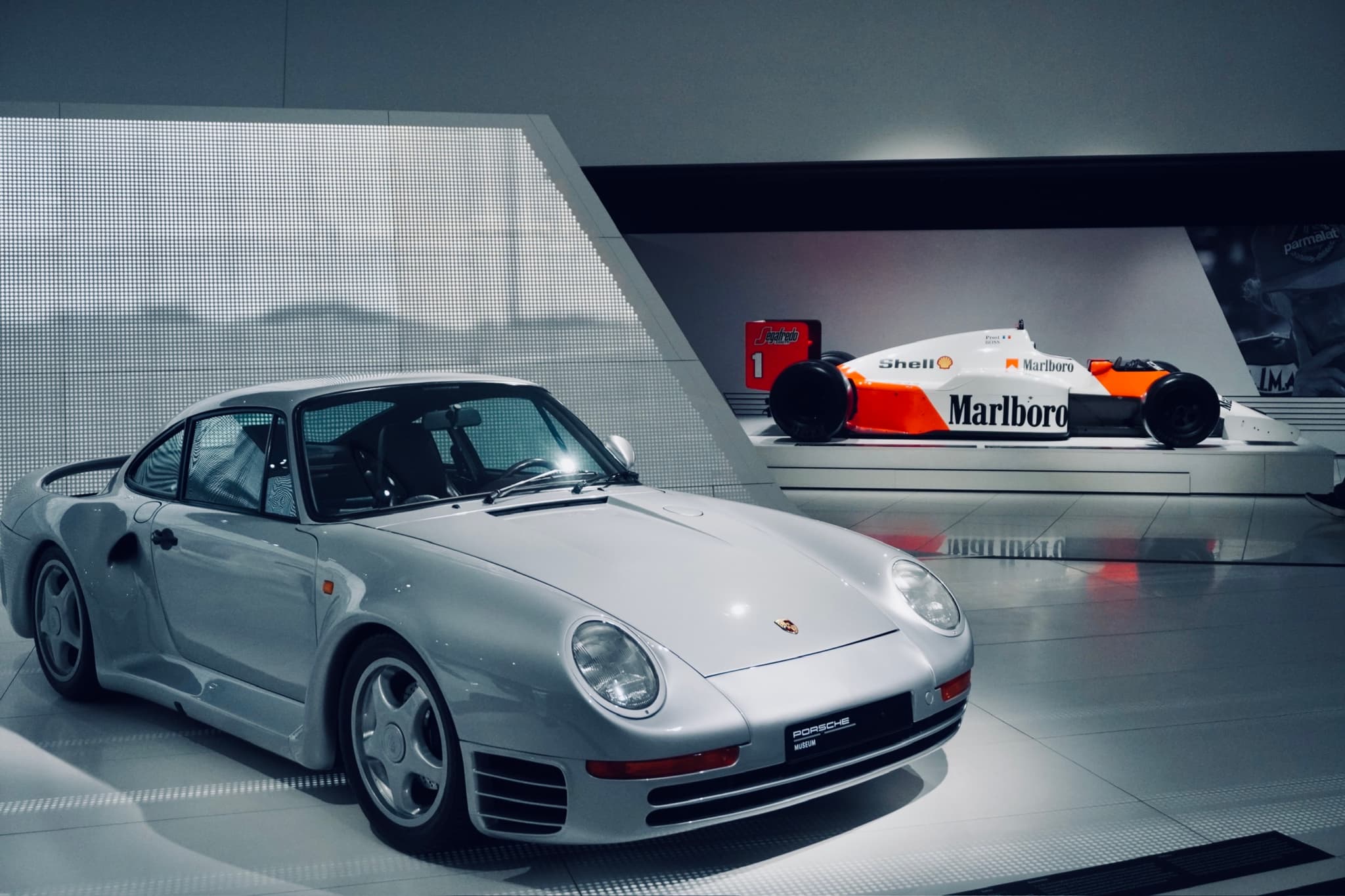 silver porsche 959 parked in a bright museum gallery with a formula one race car displayed on a raised platform behind it