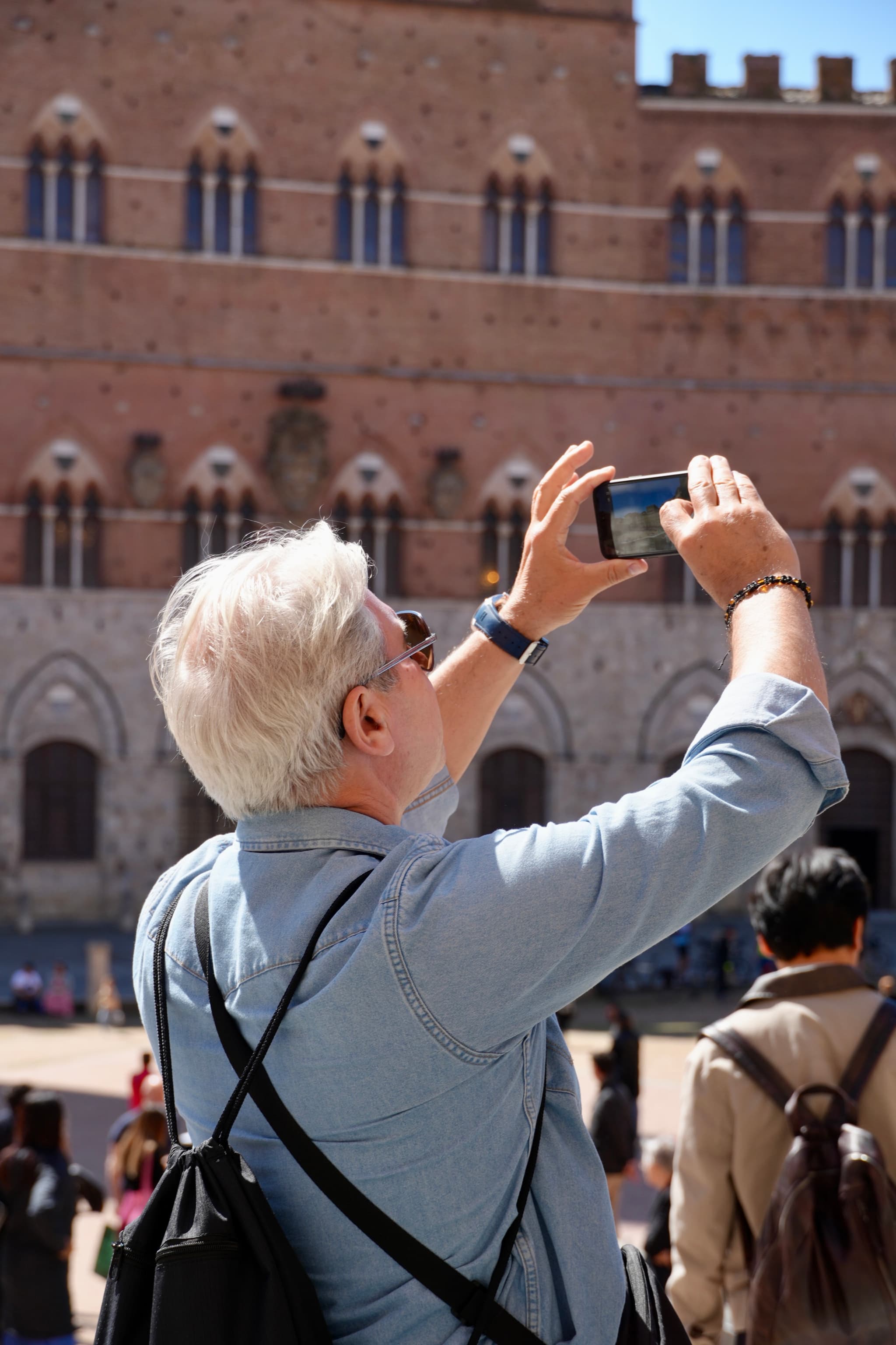 tourist with white hair uses a smartphone to photograph the red brick facade of a historic building in siena under bright sunlight