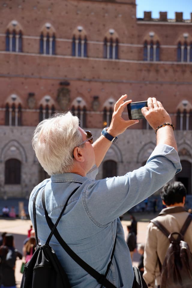 tourist with white hair uses a smartphone to photograph the red brick facade of a historic building in siena under bright sunlight