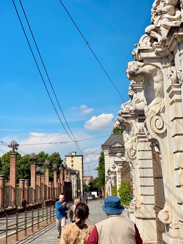people walk past ornate stone carvings on a sunny day in turin, italy, under a clear blue sky with overhead tram wires