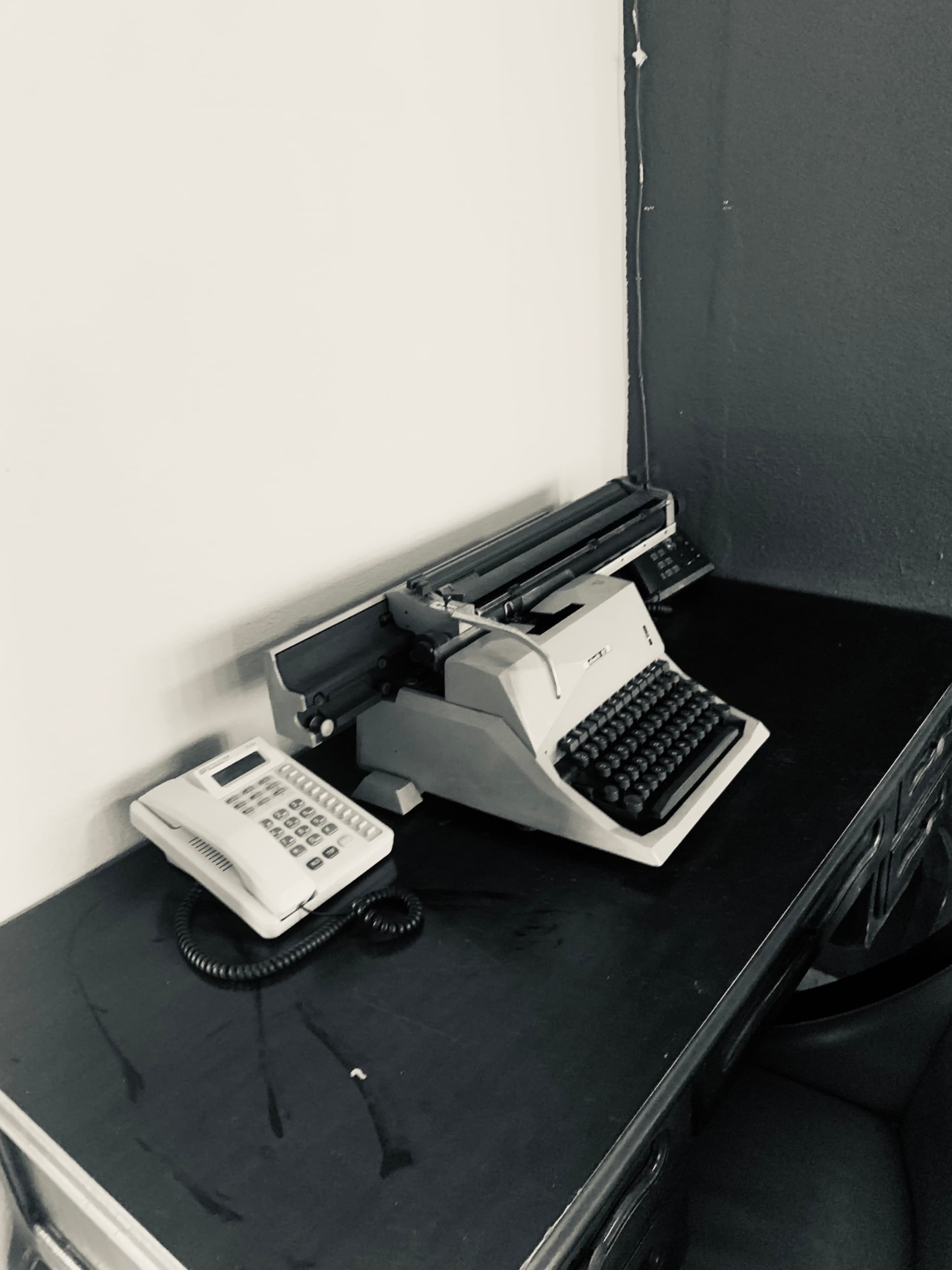 vintage typewriter and landline phone sit on a black desk against a white wall in a quiet office with overhead lighting