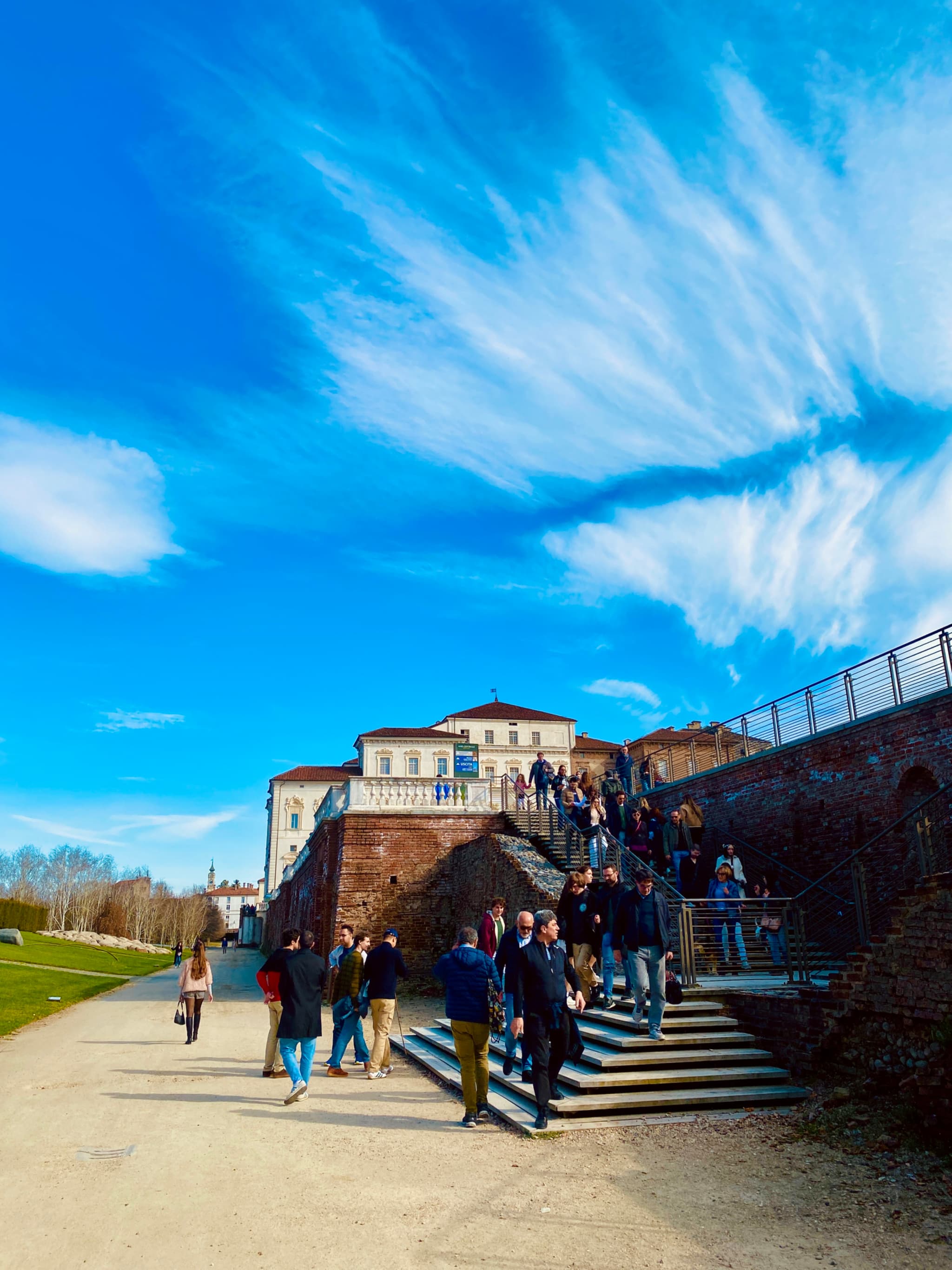 people walk down stone stairs toward a large villa under a bright blue sky with thin clouds on a sunny day
