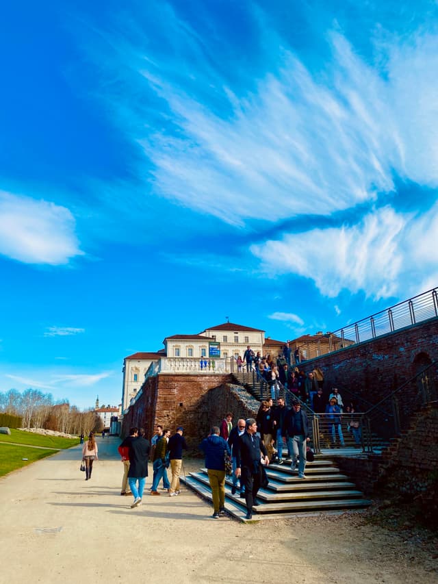 people walk down stone stairs toward a large villa under a bright blue sky with thin clouds on a sunny day
