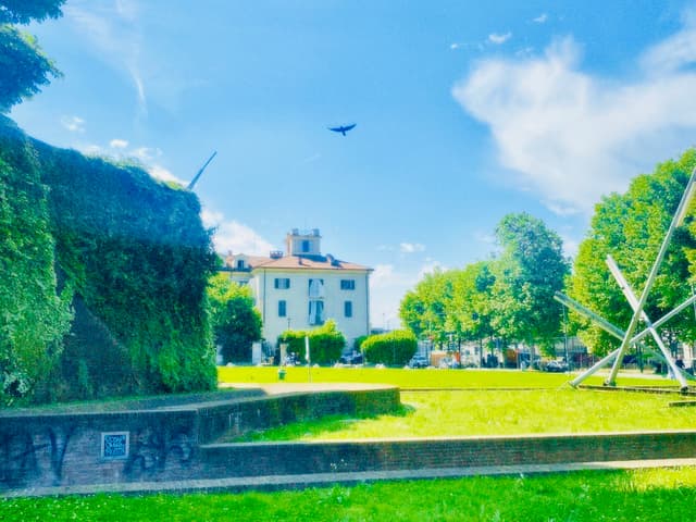 a bird flies over a sunny park with green grass, a stone wall, and a white building under a bright blue sky