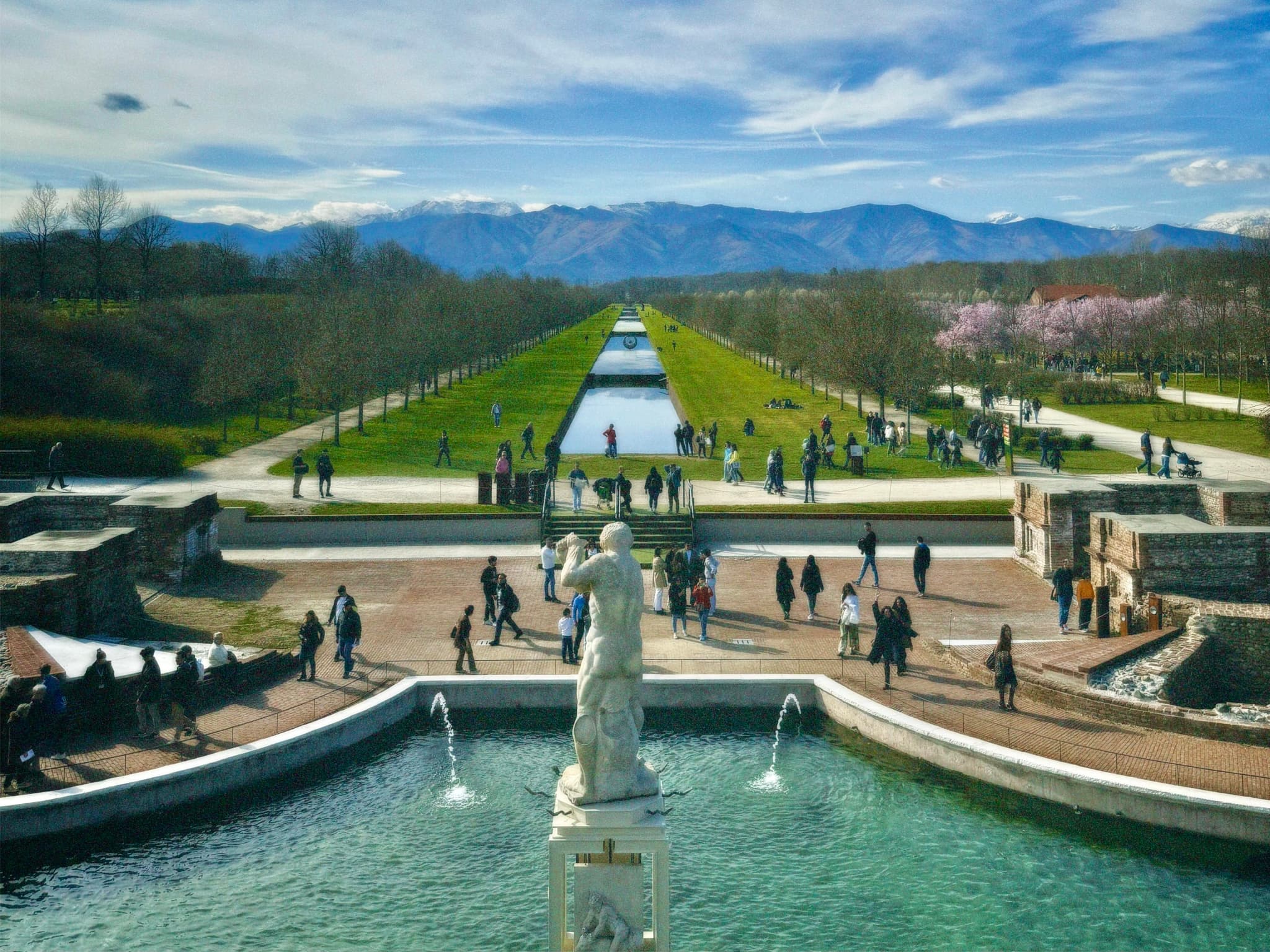 visitors walk through the palace gardens at venaria reale in italy under bright daylight with a central fountain and mountain backdrop