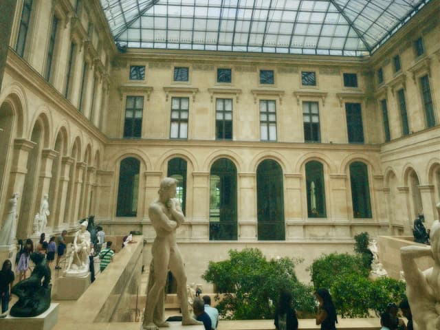 visitors walk through the louvre museum sculpture gallery under a glass ceiling with natural daylight illuminating stone statues and green indoor plants