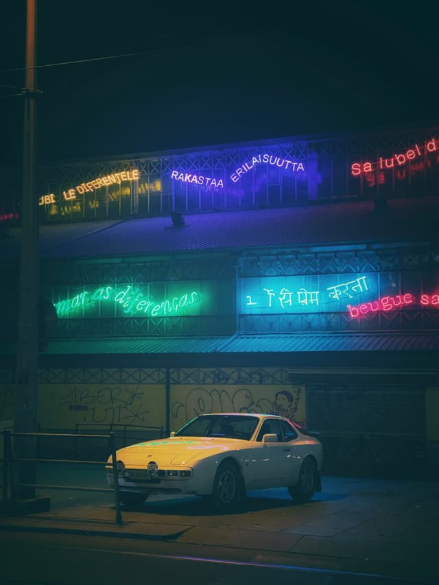 white sports car parked at night under glowing neon signs with multilingual text on a dark urban street in low light