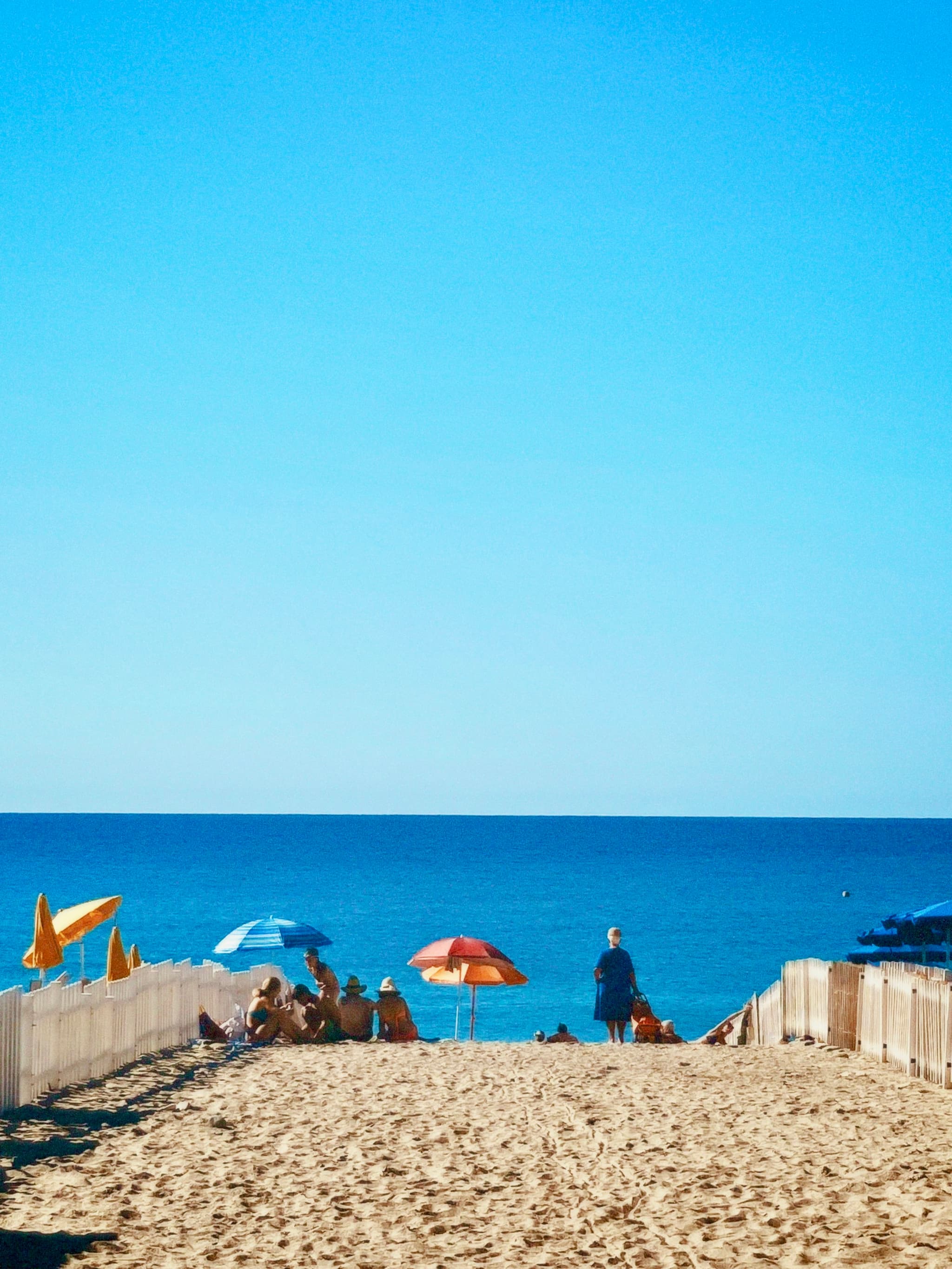 people sit under colorful umbrellas on a sandy beach path leading to the blue ocean under a clear, bright midday summer sky