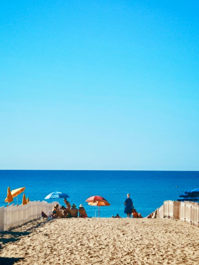 people sit under colorful umbrellas on a sandy beach path leading to the blue ocean under a clear, bright midday summer sky