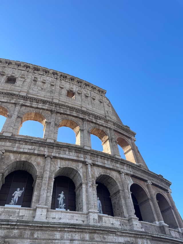 low angle view of the stone colosseum in rome under a clear blue sky with bright sunlight highlighting the ancient arched facade