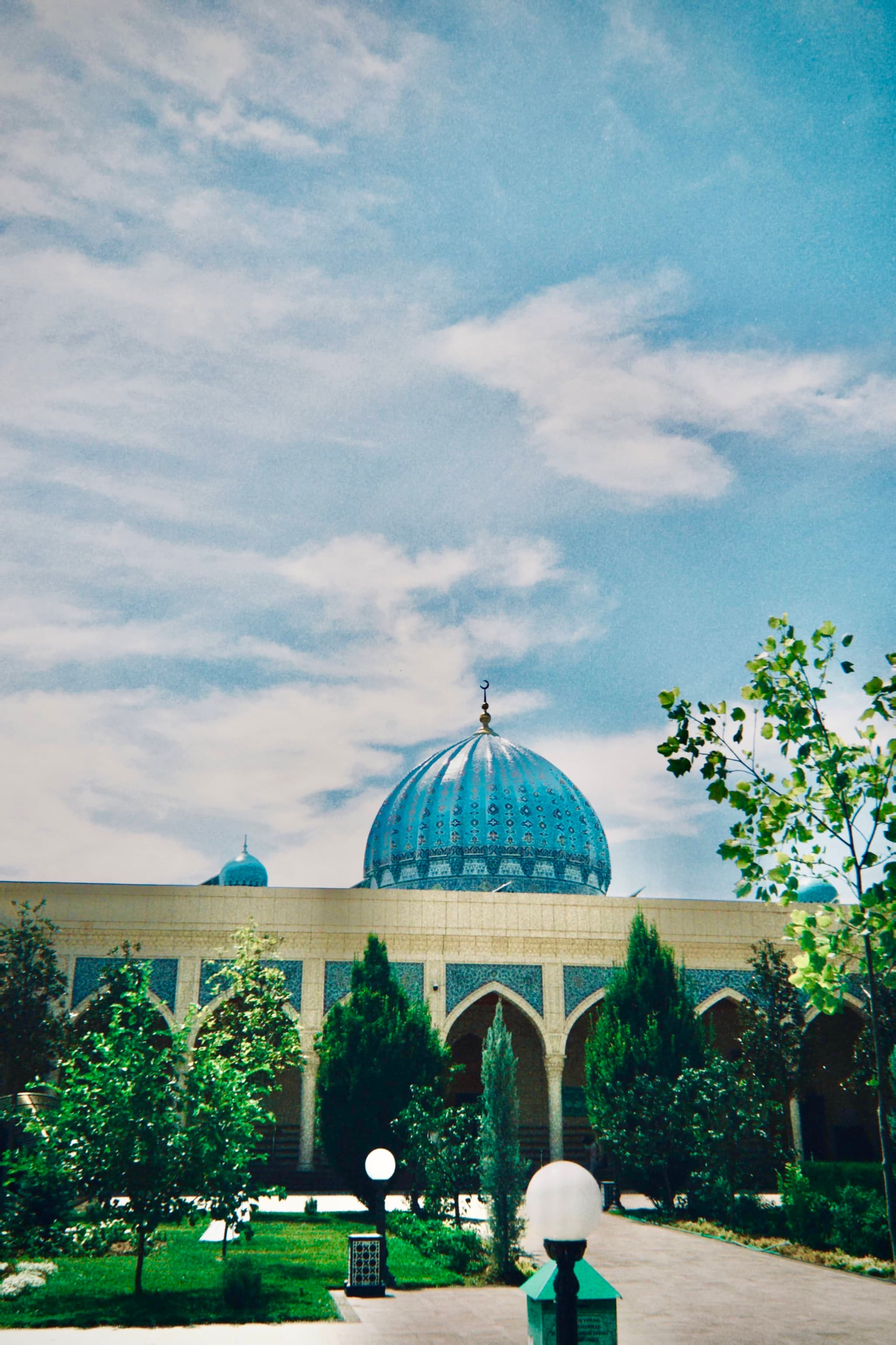 blue dome of a mosque rises above green trees and arched walkways under a bright sky with scattered clouds in daylight