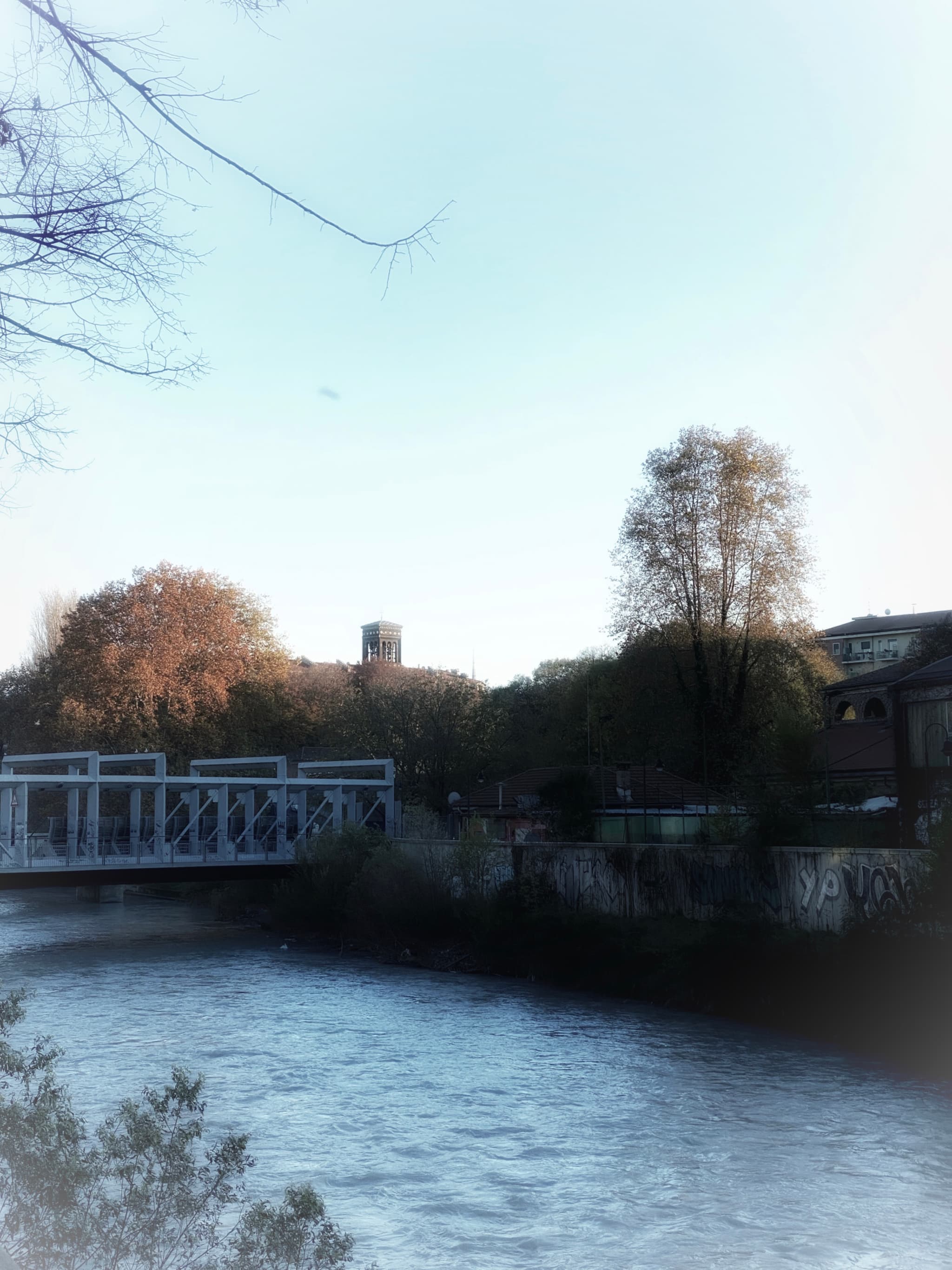 metal footbridge crosses a river near trees and a distant water tower under clear daylight in a quiet urban park setting
