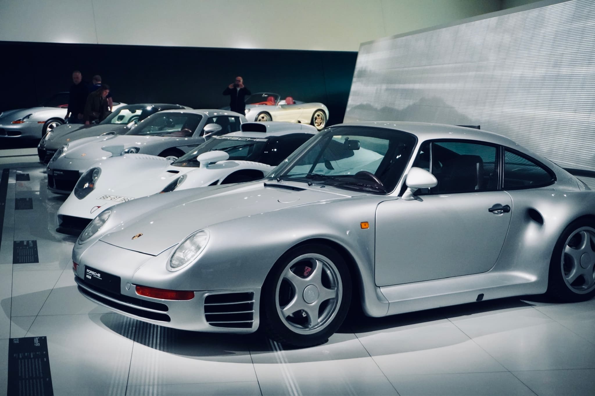 silver porsche sports cars parked on a white floor inside a modern museum under bright overhead lights with visitors in background