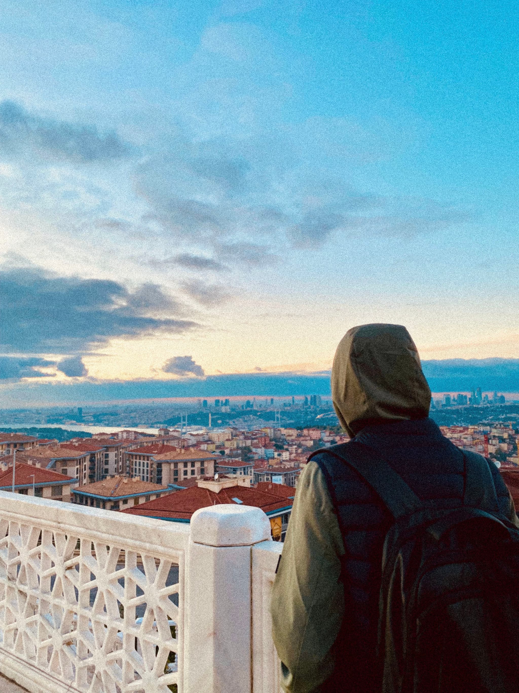 person in a hooded jacket looks over the istanbul skyline from a stone balcony during a clear sunset with soft light