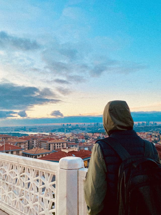 person in a hooded jacket looks over the istanbul skyline from a stone balcony during a clear sunset with soft light