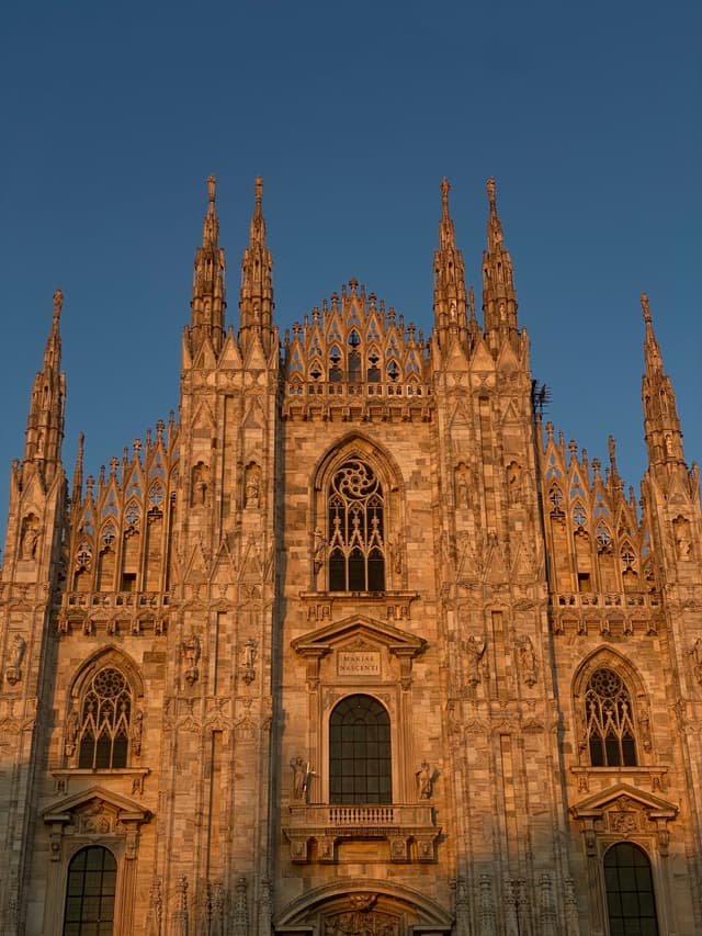 milan cathedral facade lit by warm sunset light against a clear blue sky, showing detailed gothic spires and stone carvings in italy
