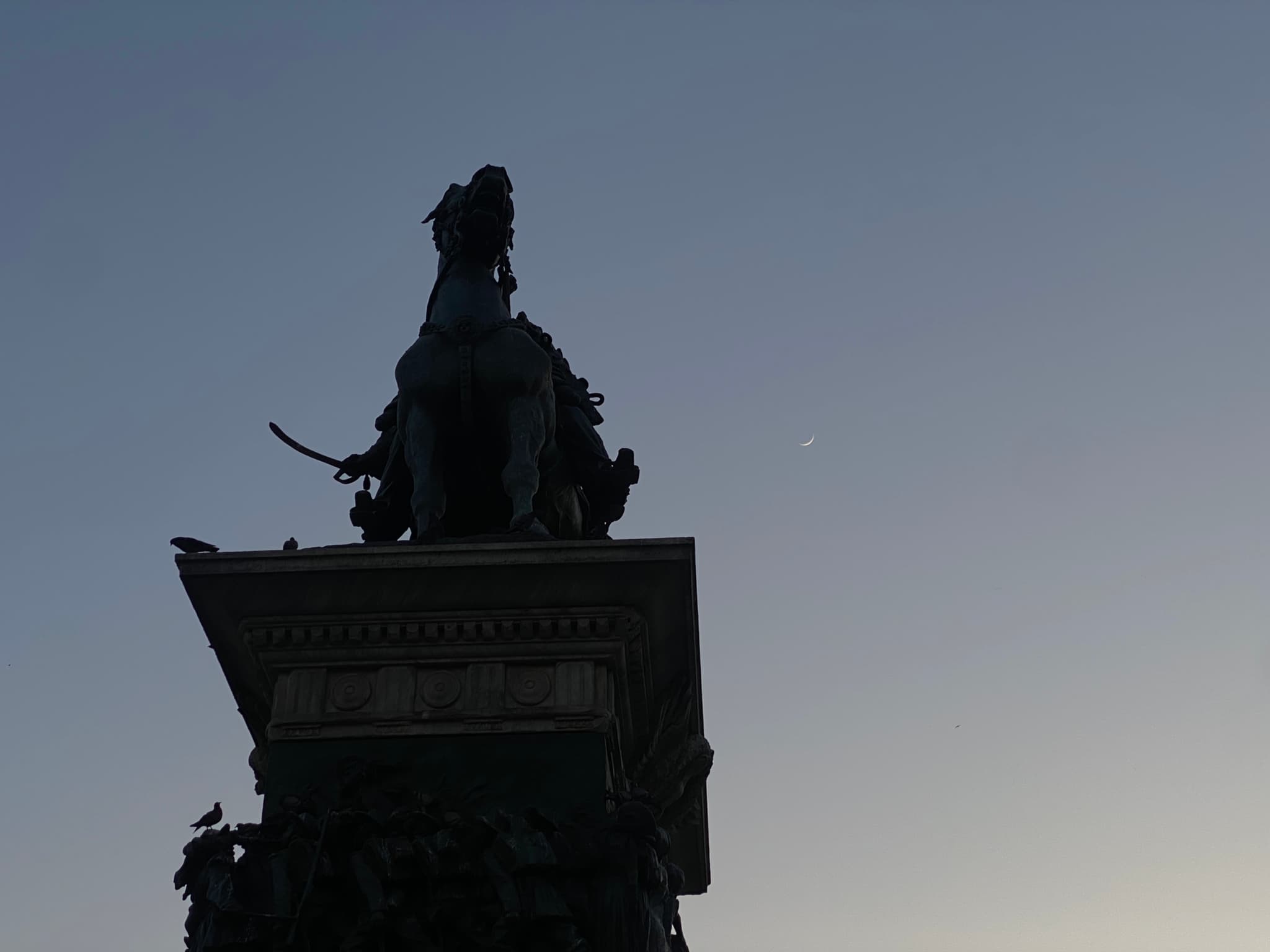 silhouette of a bronze equestrian statue against a clear evening sky with a visible moon in a quiet public square setting