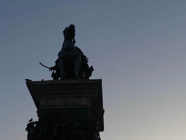 silhouette of a bronze equestrian statue against a clear evening sky with a visible moon in a quiet public square setting