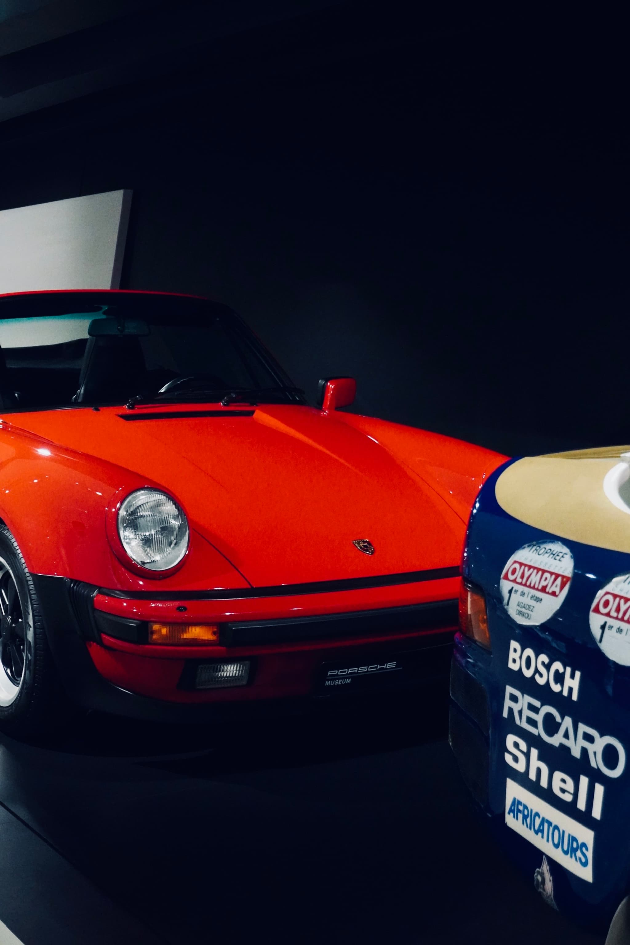 bright red porsche 911 parked next to a race car in a dark museum gallery with focused overhead lighting and clean lines