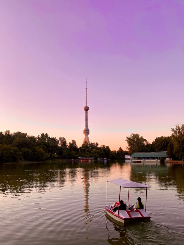 people ride a pedal boat on a calm lake during sunset with the tall tashkent television tower visible in the background