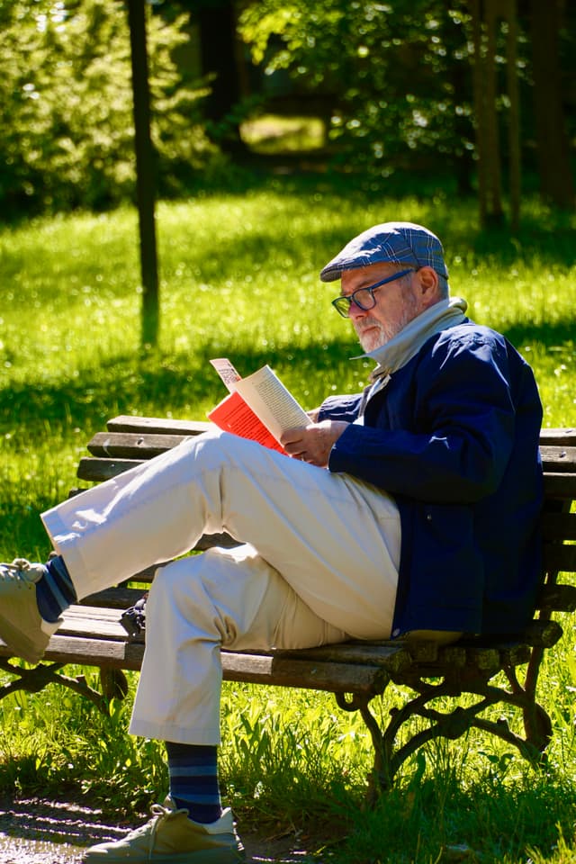 elderly man in a flat cap reads a book on a wooden park bench during a bright afternoon with dappled sunlight