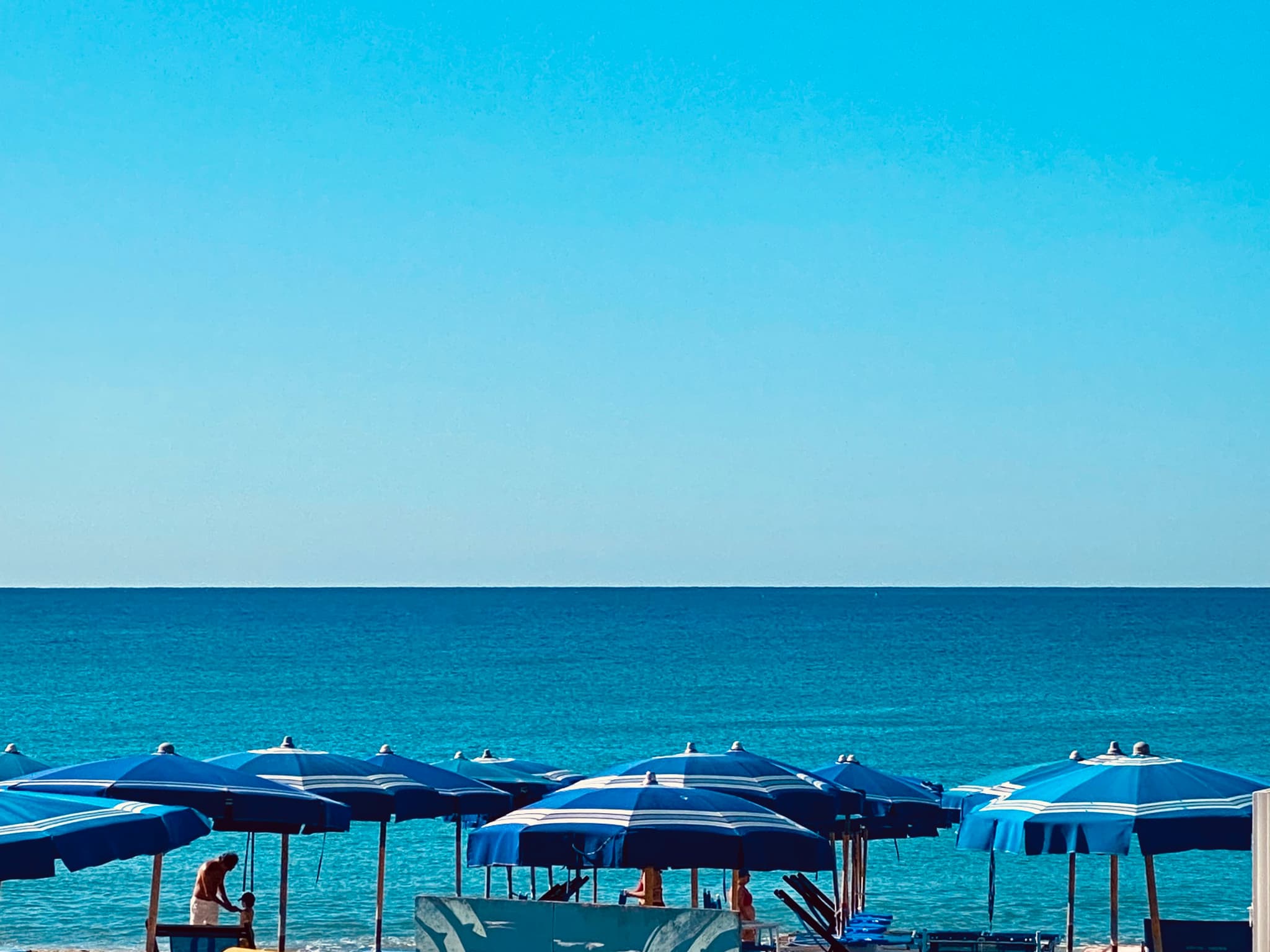 blue striped umbrellas line a sunny beach under a clear sky, overlooking the calm sea on a bright summer afternoon in italy