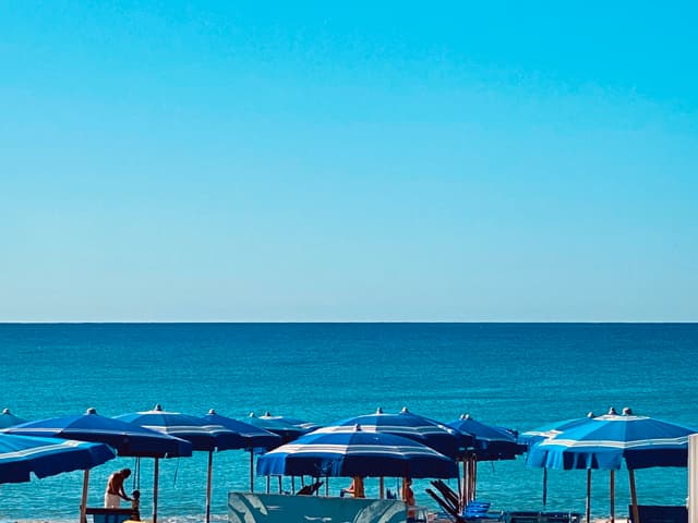 blue striped umbrellas line a sunny beach under a clear sky, overlooking the calm sea on a bright summer afternoon in italy