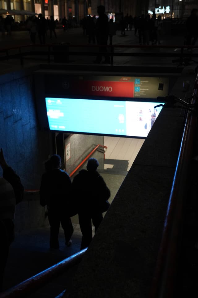 commuters walk down stairs into a bright subway entrance at night, illuminated by a glowing rectangular sign under dark city skies