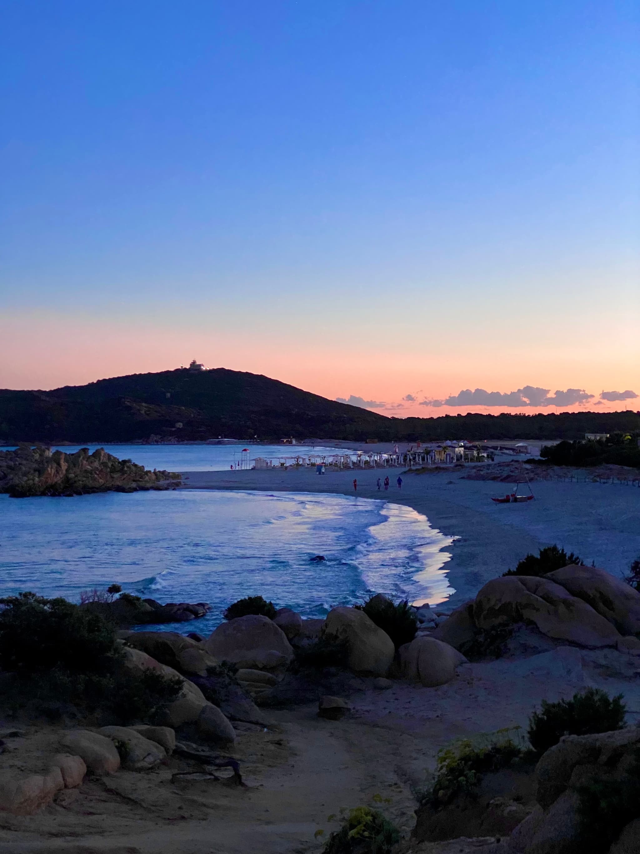 sandy beach at sunset with blue shadows, granite rocks in the foreground, and a distant hill under a clear evening sky in sardinia